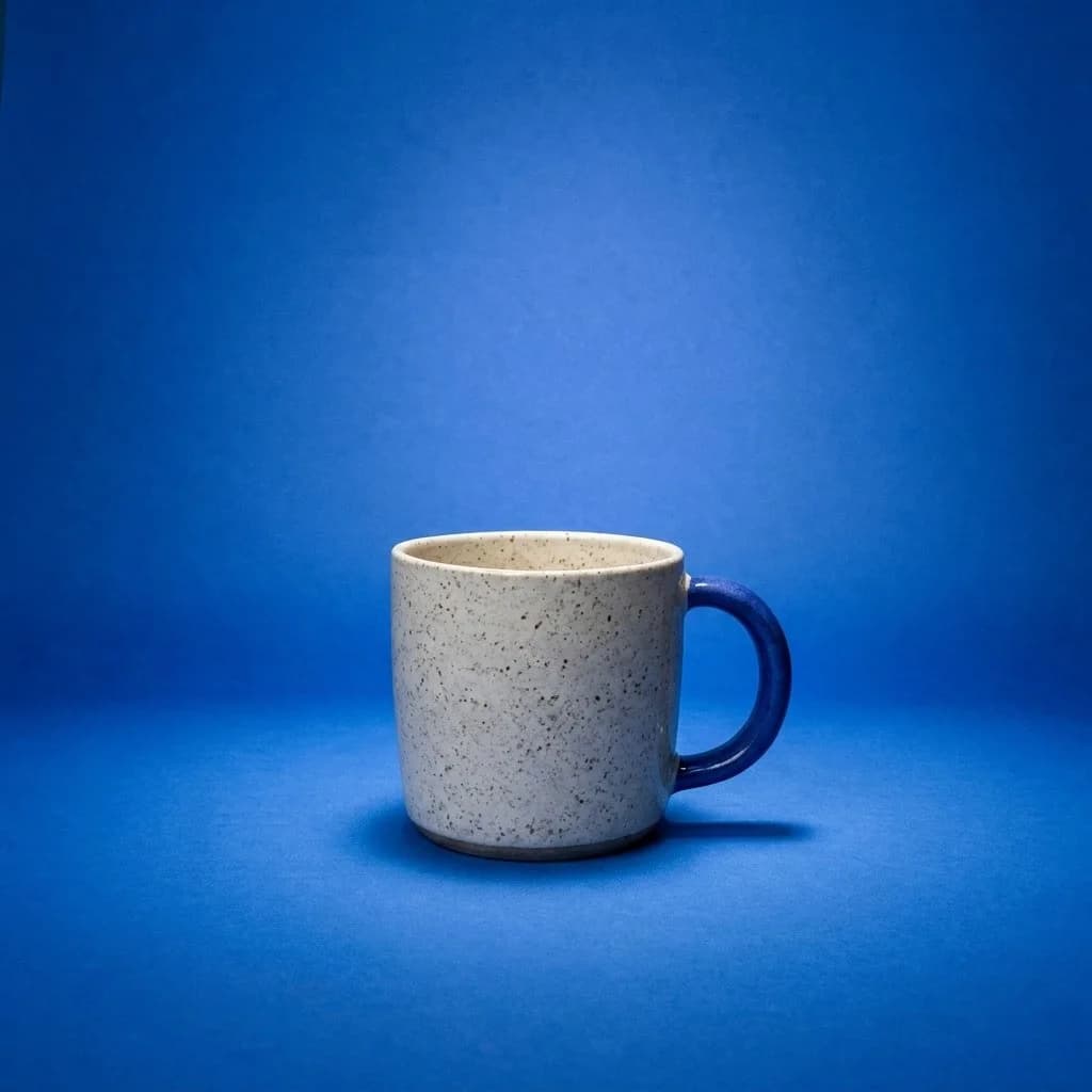 Taza artesanal crema moteada con asa azul sobre fondo azul cobalto y un foco de luz suave.