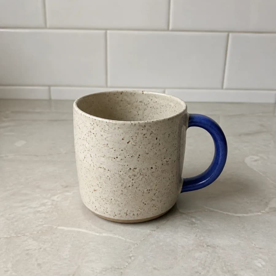 Taza artesanal de cerámica crema moteada con asa azul fotografiada en una cocina.